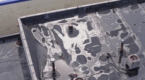 Standing water and pooling on a flat roof membrane around vents and parapet walls.