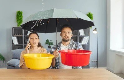 Worried couple sitting under an umbrella indoors catching ceiling roof leak water in buckets.