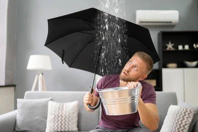 Man sitting on couch holding umbrella and bucket under rain leaking from ceiling after storm,