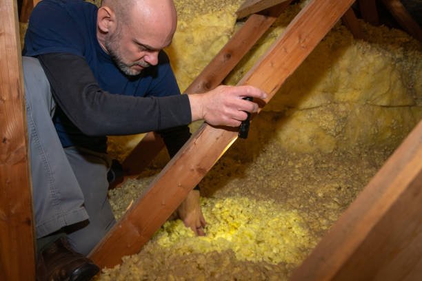 Roofing contractor using a light to examine wooden roof rafters and fiberglass insulation in a New Jersey attic.