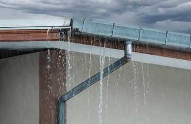 Water overflowing from a clogged roof gutter and downspout system during rain damage inspection.