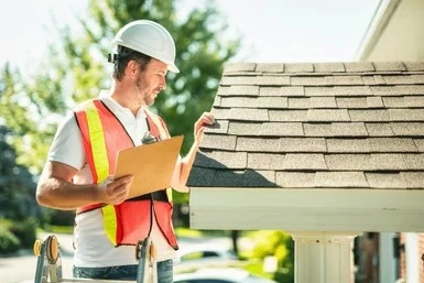 Roof inspector in a hard hat and safety vest holding a clipboard during a residential roof inspection.
